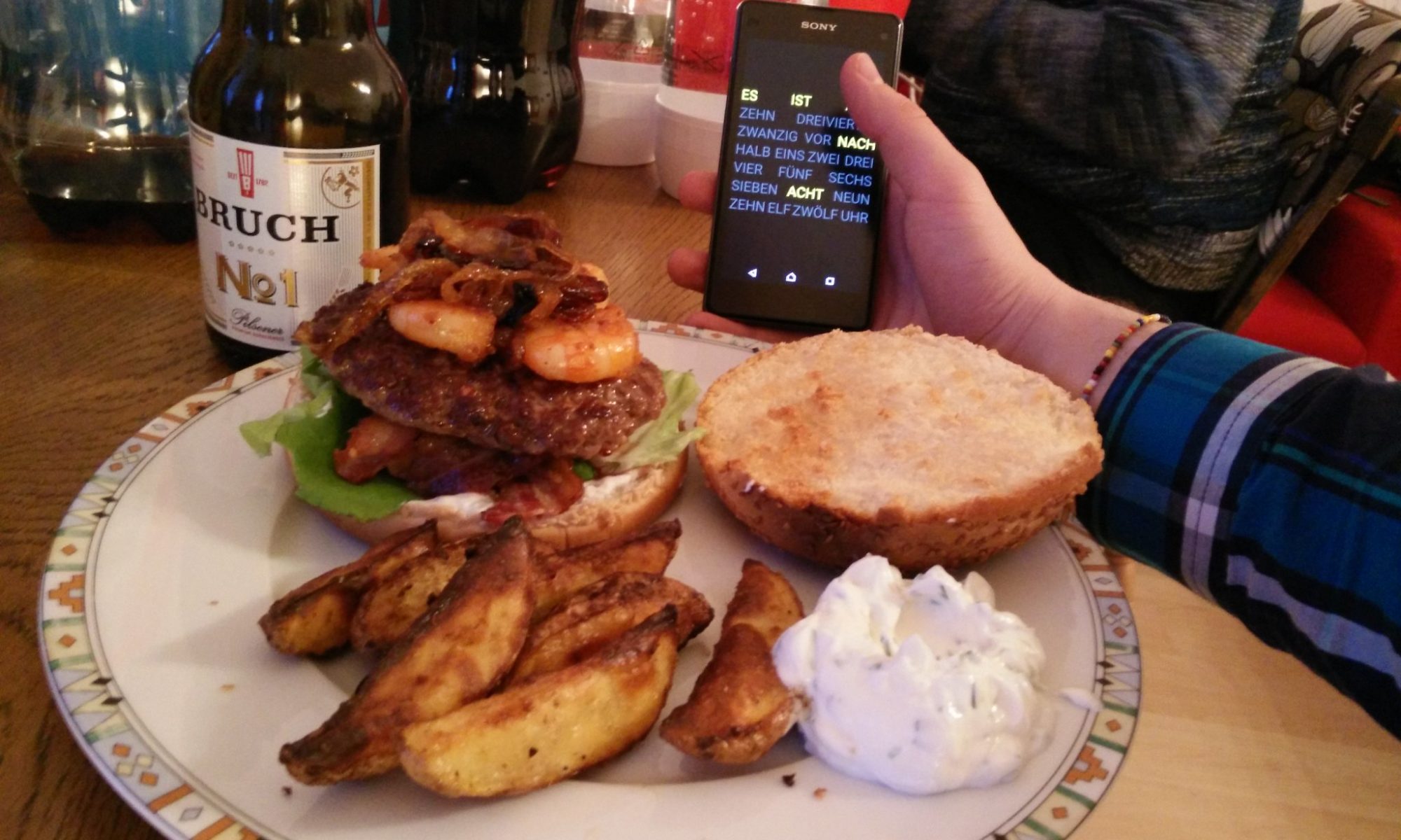 Ein Teller mit einem Burger, Kartoffelecken und Soße, platziert auf einem Tisch mit Bier und dabei ein Smartphone gehalten von einer Hand und mit der Wortuhr als Displayanzeige
